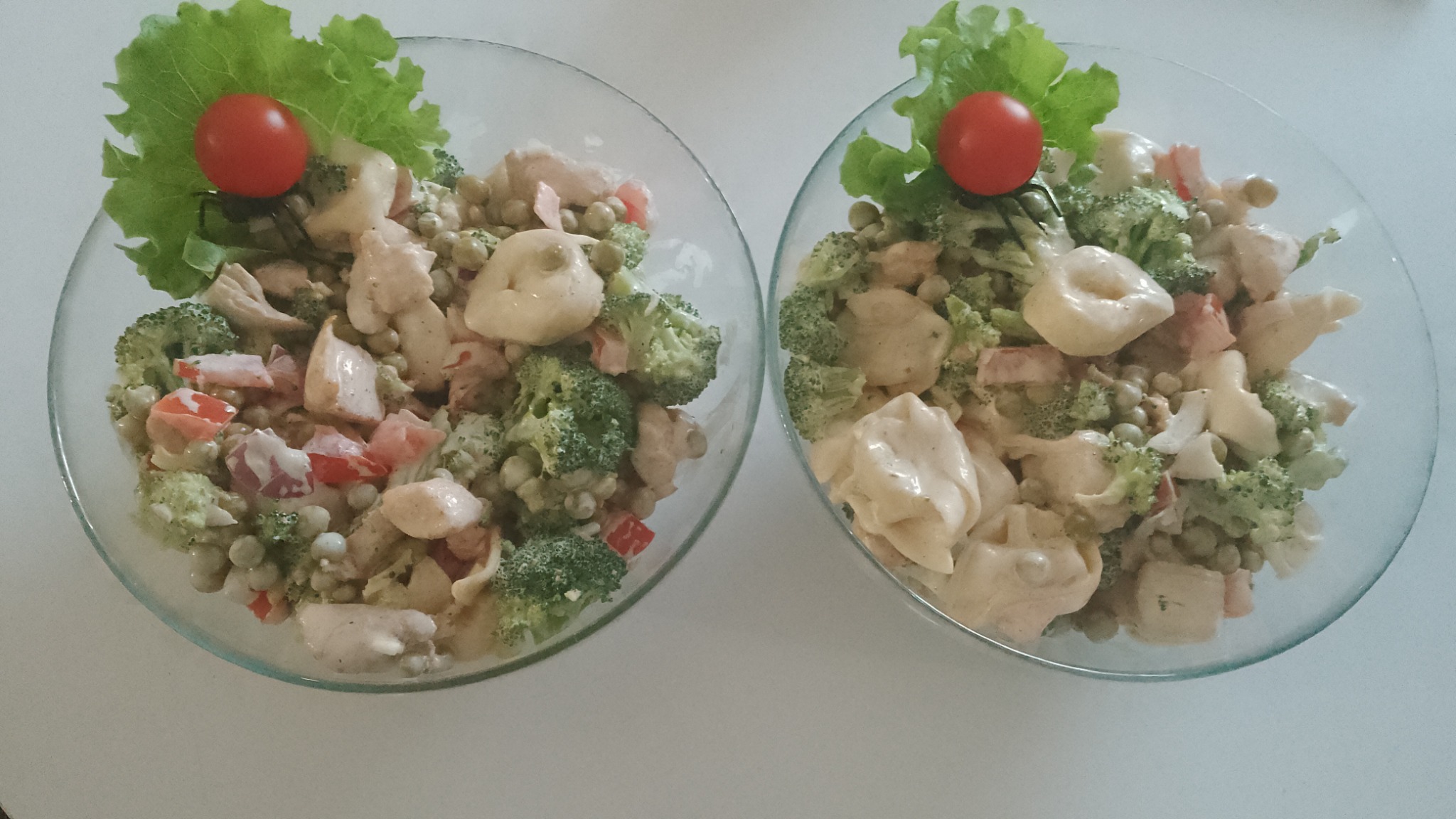 Dwie szklane miski z sałatką: brokuły, groszek, pierożki tortellini, kurczak i pomidor, udekorowane liściem sałaty i pomidorkiem cherry na białym tle.