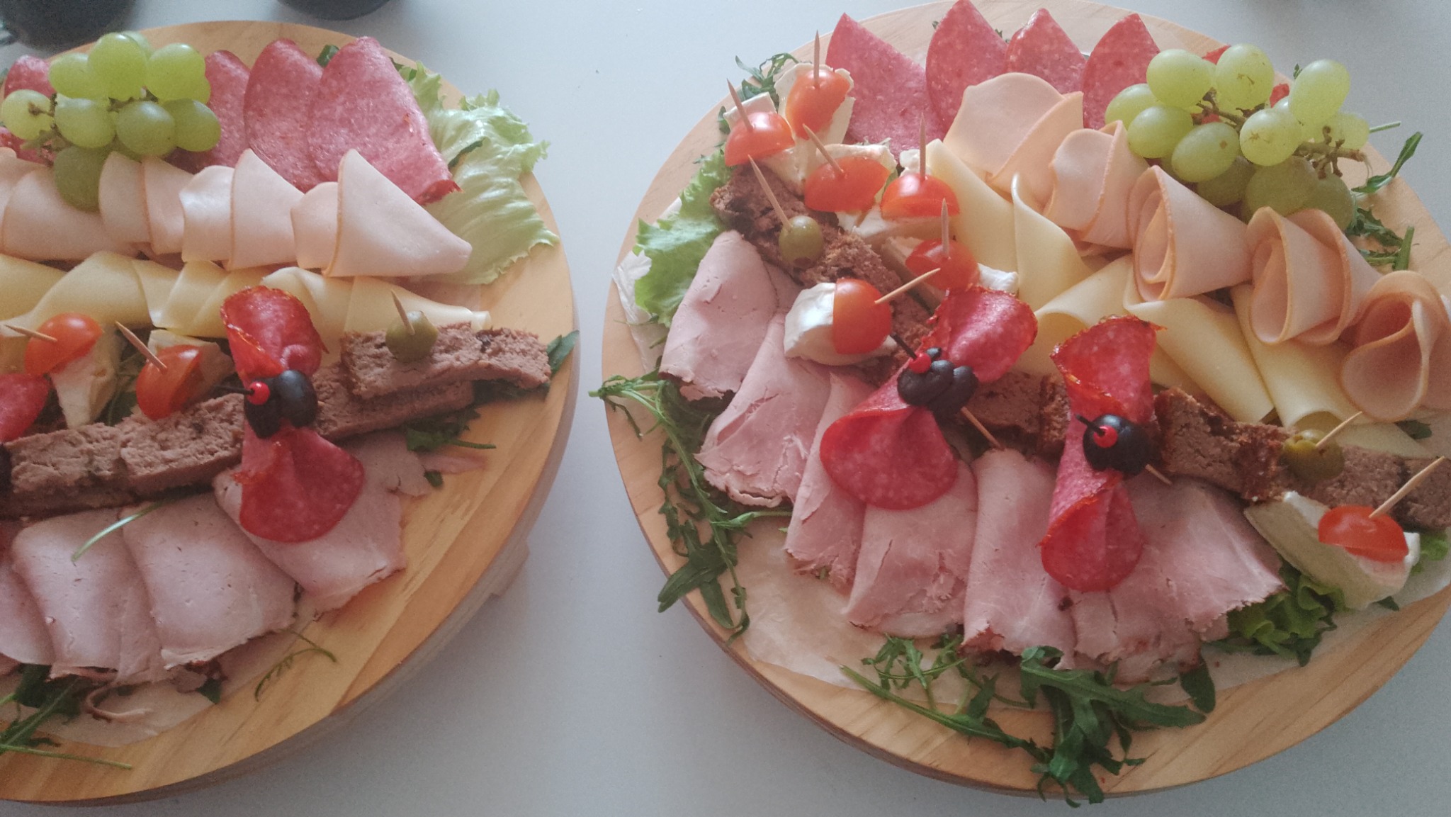 Dwa drewniane talerze z różnorodnymi przekąskami: wędliny, sery, pieczywo, winogrona, pomidorki koktajlowe i zielenina, ułożone dekoracyjnie na białym tle.