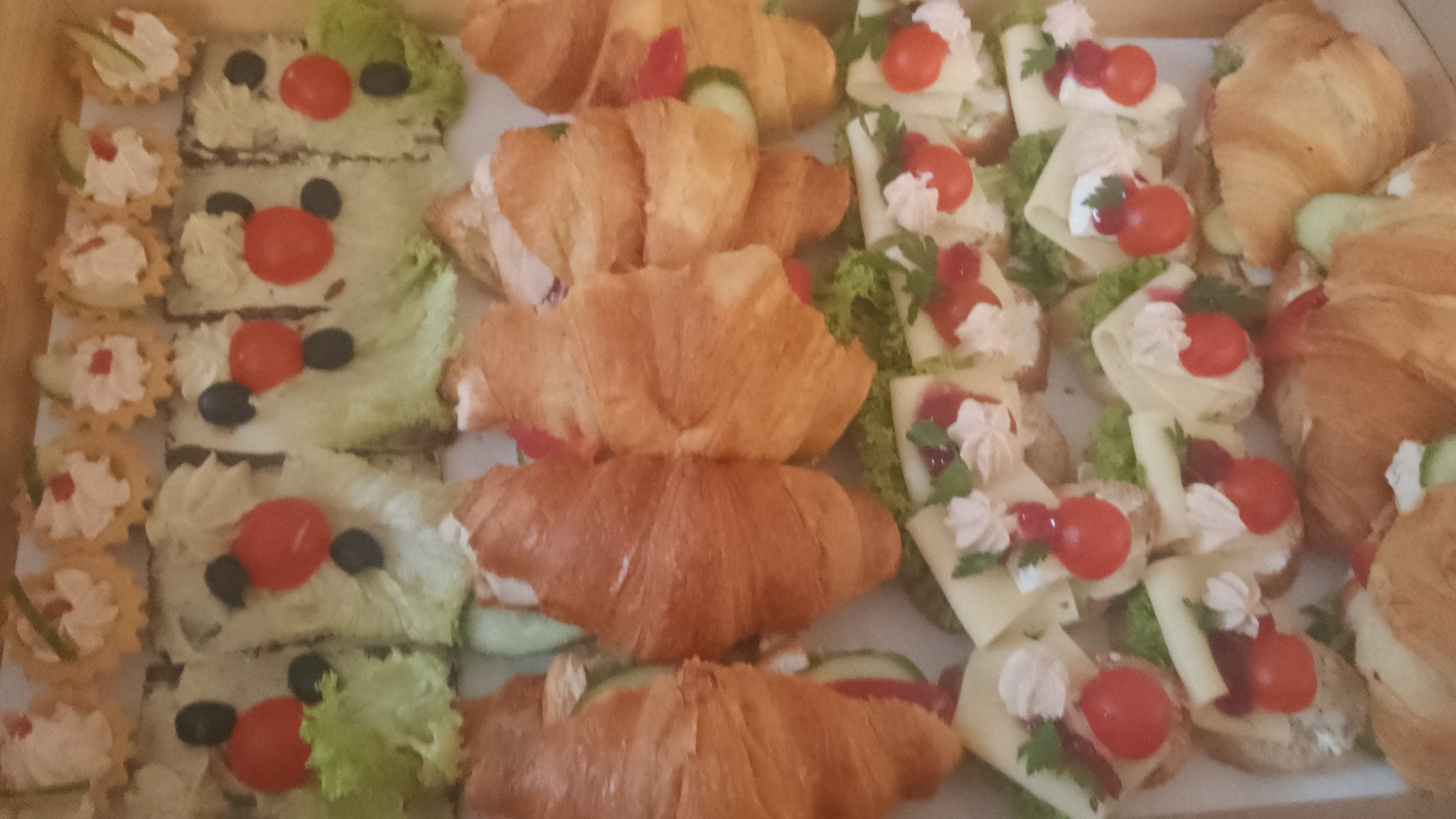 Różnorodne przekąski w kartonowym pudełku: mini tartaletki z kremem, kanapki z warzywami ułożone w kształt klauna oraz croissanty z nadzieniem.