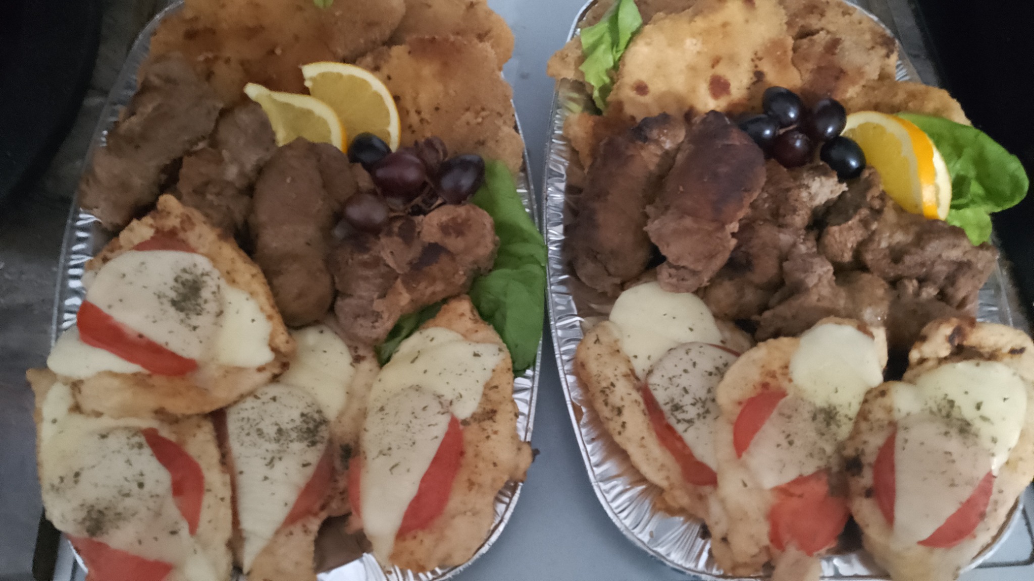 Dwa aluminiowe półmiski z różnorodnymi daniami mięsnymi: kotlety schabowe, pieczeń, grillowane kiełbaski, kurczak zapiekany z serem i pomidorem, dekorowane sałatą, winogronami i plasterkiem...