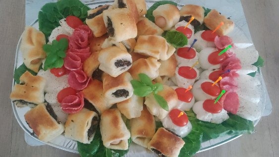 Elegancki półmisek z przekąskami: roladki z ciasta francuskiego z farszem, koreczki z mozzarelli i pomidorków, dekoracje z salami, całość na liściach sałaty.