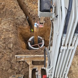 Wiesław Markowicz - Głęboki wykop w ziemi z betonową studnią kanalizacyjną opuszczaną do środka za pomocą łańcucha przez maszynę wykopową z widocznymi przewodami hydraulicznymi.