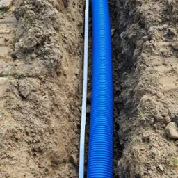 Niebieska rura karbowana ułożona w wąskim wykopie w ziemi, obok leży metalowa listwa pomiarowa.