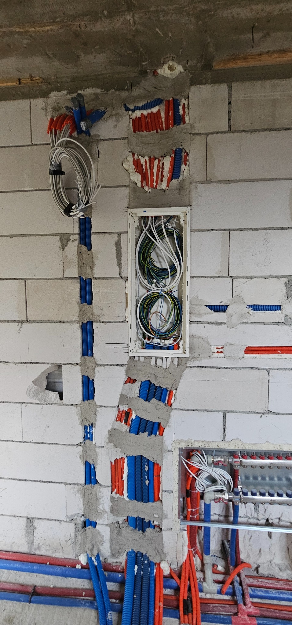 Ściana z betonu komórkowego z bruzdami wypełnionymi instalacjami w peszlach, widoczna skrzynka rozdzielcza z przewodami i rozdzielacz ogrzewania podłogowego.