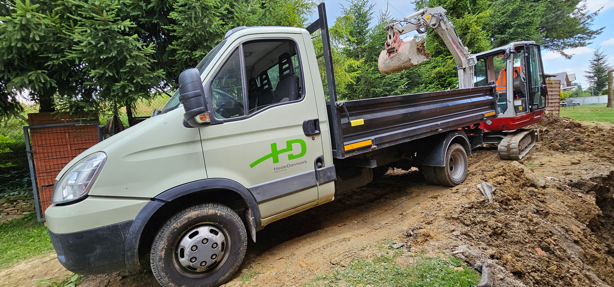 Koparka Takeuchi nabiera ziemię do wykopu obok ciężarówki HedeDanmark na terenie z drzewami iglastymi w tle.