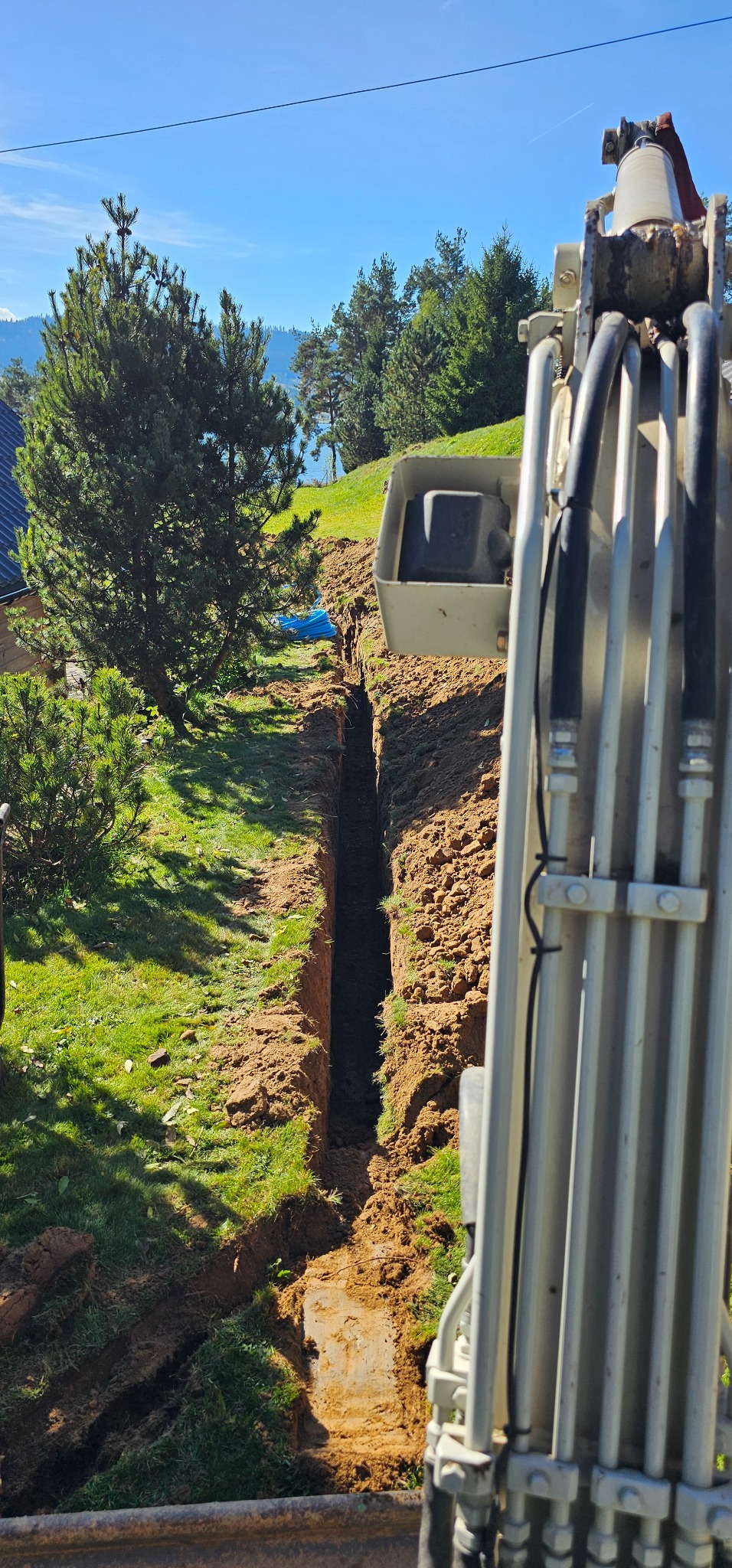 Wąski wykop liniowy w trawiastym terenie, wykonany prawdopodobnie pod instalacje, widoczny z perspektywy operatora koparki.