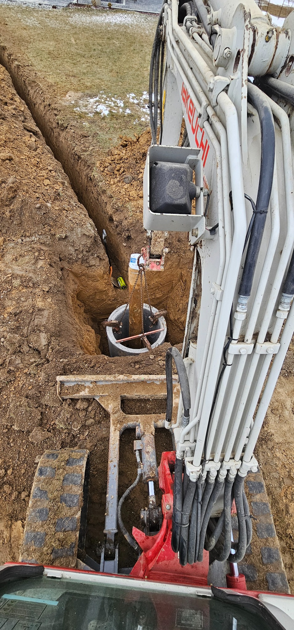 Głęboki wykop w ziemi z betonową studnią kanalizacyjną opuszczaną do środka za pomocą łańcucha przez maszynę wykopową z widocznymi przewodami hydraulicznymi.