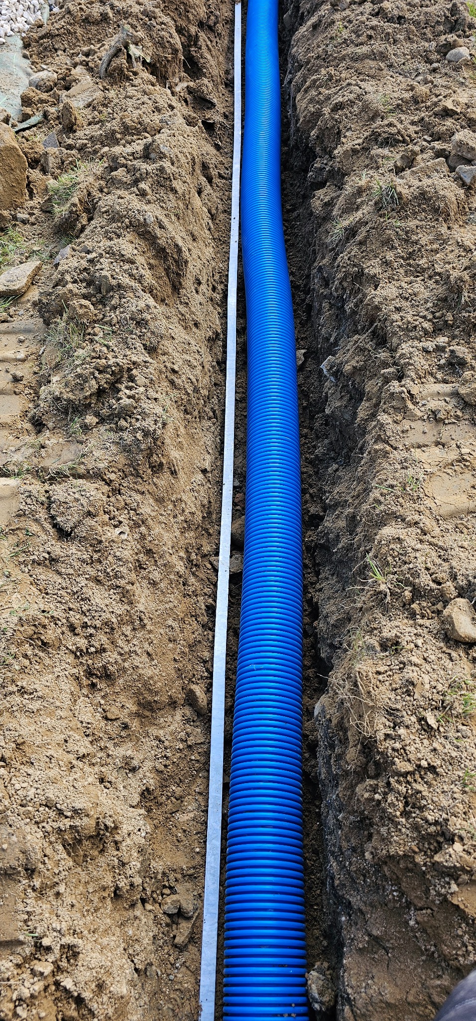 Niebieska rura karbowana ułożona w wąskim wykopie w ziemi, obok leży metalowa listwa pomiarowa.