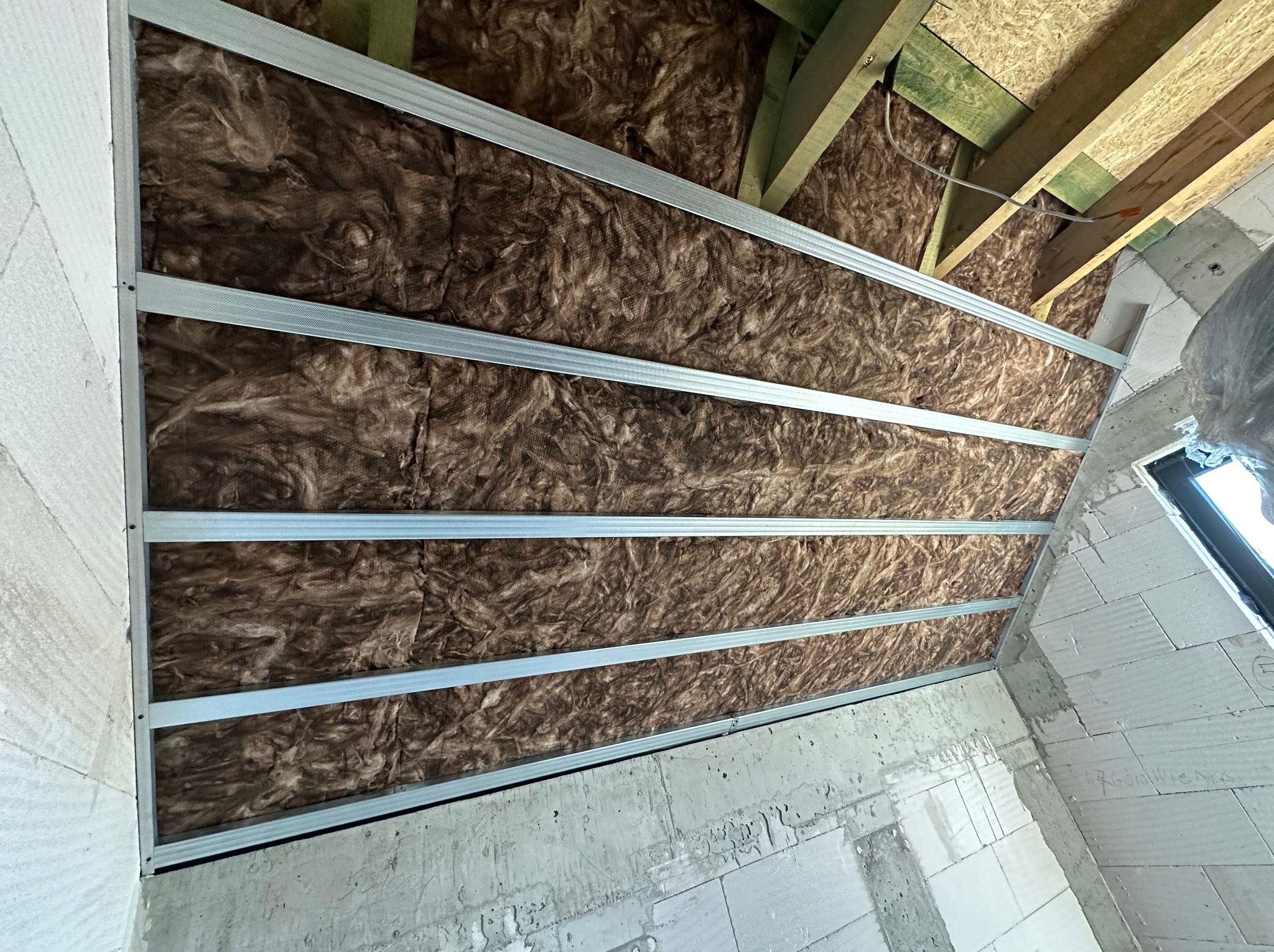 Poddasze w trakcie izolacji termicznej, widoczne brązowe wypełnienie między metalowymi profilami konstrukcyjnymi, surowe ściany z betonu komórkowego i drewniane elementy więźby dachowej.