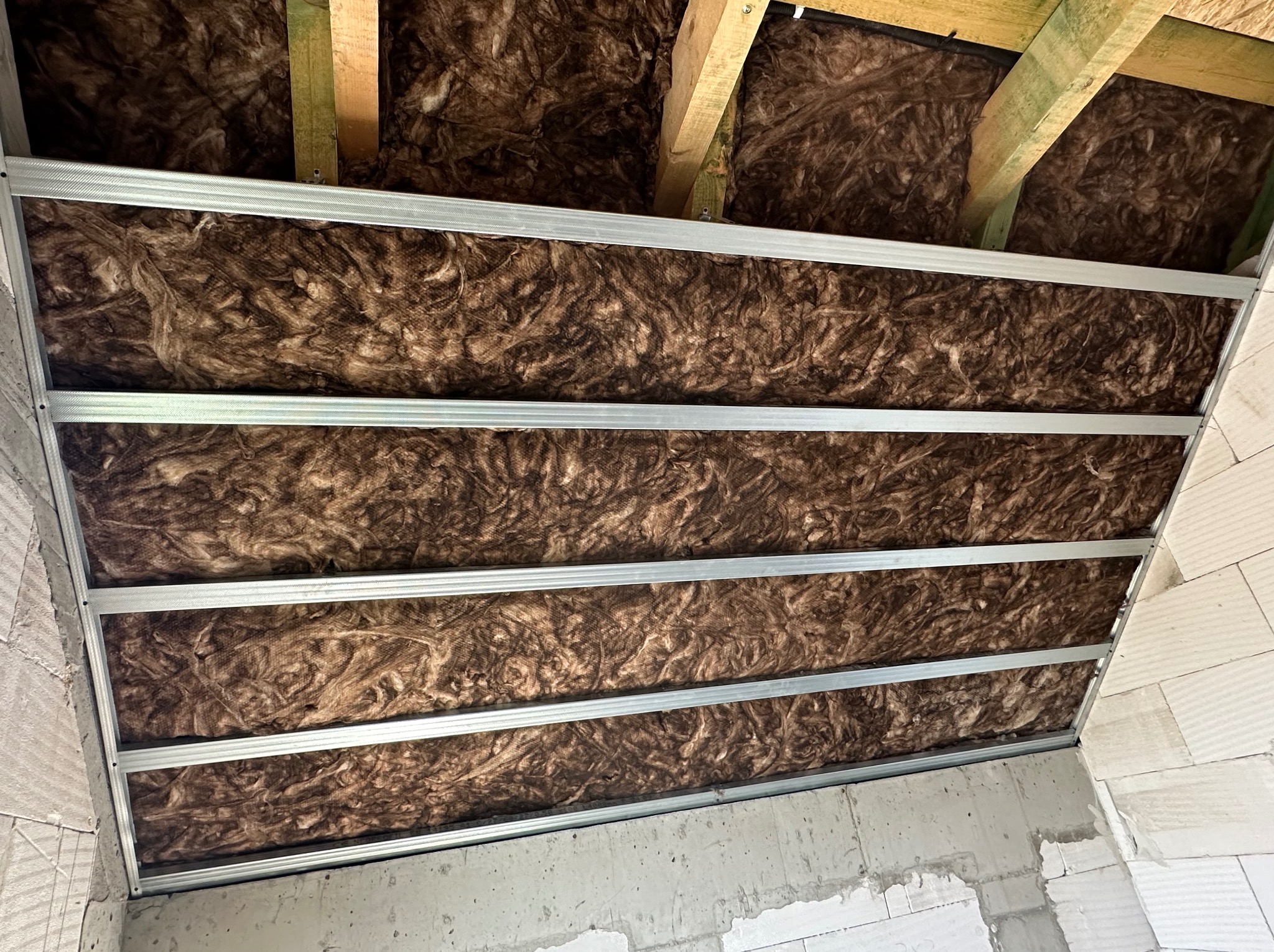 Izolacja z wełny mineralnej między krokwiami dachu, zabezpieczona metalowym stelażem, widok od dołu na surową ścianę z betonu komórkowego.