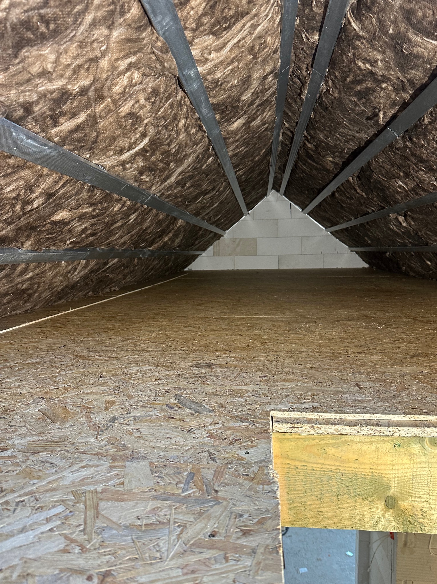 Poddasze z widoczną izolacją z wełny mineralnej między krokwiami, wykończone płytą OSB, z fragmentem ściany z bloczków betonowych w głębi.