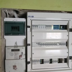 VOLT GREMIUM KAROL MICHAŁEK - Zamontowana skrzynka rozdzielcza z bezpiecznikami, obok licznik energii elektrycznej Norax 3 w surowym wnętrzu z widocznymi przewodami. Widoczne elementy instalacji elektrycznej.