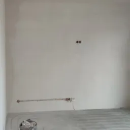 Ściana w trakcie remontu z widocznymi przewodami elektrycznymi w bruzdach, puszkami instalacyjnymi i ogrzewaniem podłogowym w wylewce. Wiadro z resztkami materiałów budowlanych w rogu pomieszczenia.