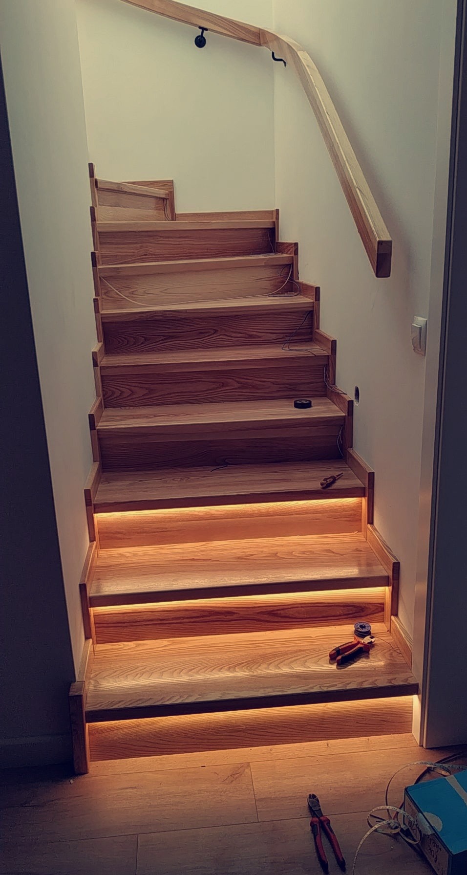Drewniane schody z podświetleniem LED na stopniach, widoczne narzędzia elektryczne i przewody, porządek prac w trakcie realizacji.