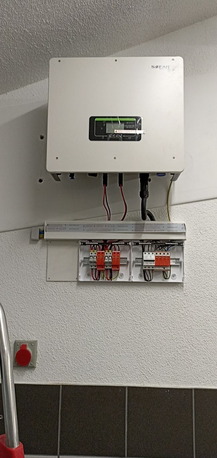 Biały falownik firmy SOFAR zamontowany na ścianie nad rozdzielnicą elektryczną z zabezpieczeniami przeciwprzepięciowymi, widoczny fragment czerwonego wtyku siłowego i fragment wózka transportowego.