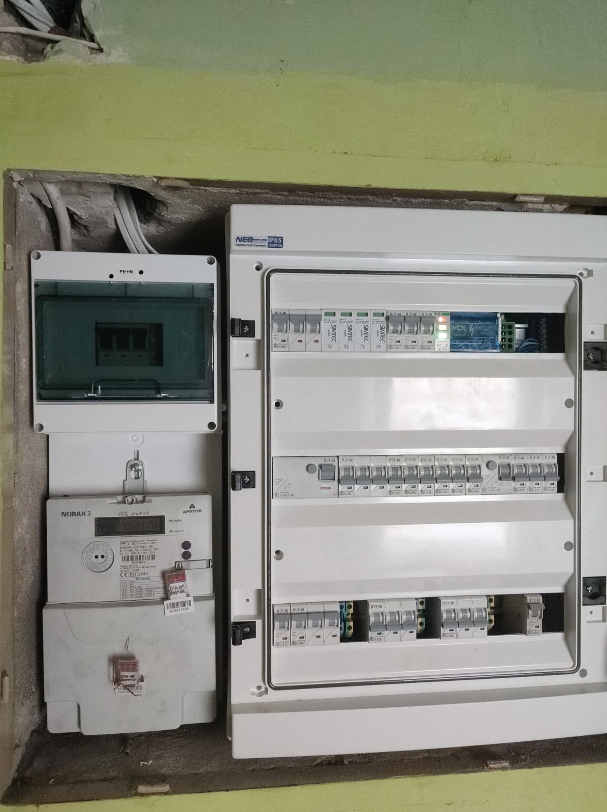 Zamontowana skrzynka rozdzielcza z bezpiecznikami, obok licznik energii elektrycznej Norax 3 w surowym wnętrzu z widocznymi przewodami. Widoczne elementy instalacji elektrycznej.