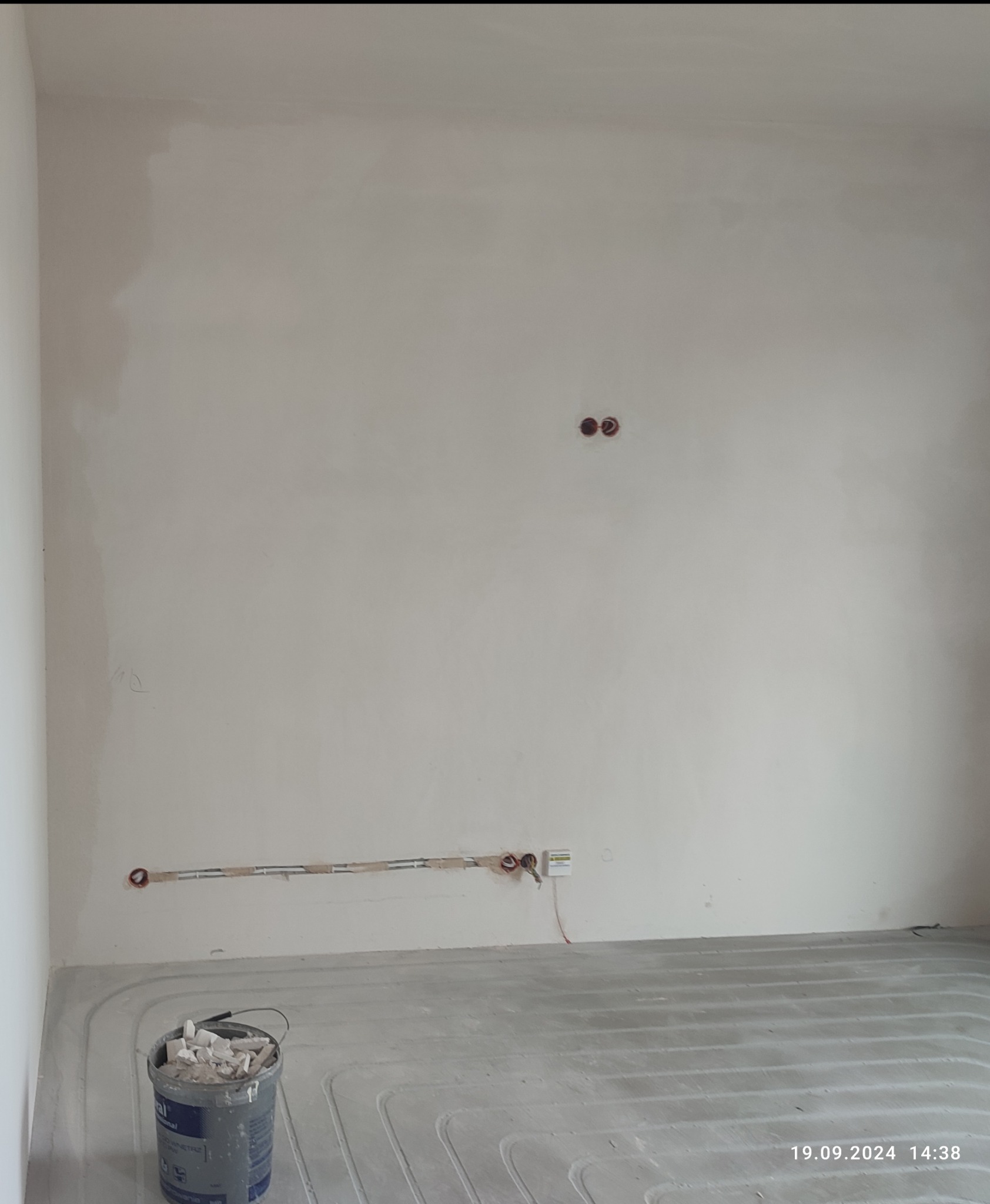 Ściana w trakcie remontu z widocznymi przewodami elektrycznymi w bruzdach, puszkami instalacyjnymi i ogrzewaniem podłogowym w wylewce. Wiadro z resztkami materiałów budowlanych w rogu pomieszczenia.