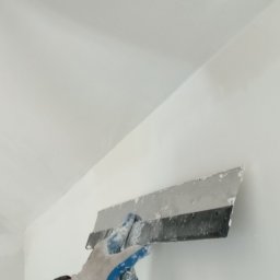 SKAWIŃSCY PROJEKTOWANIE I OBSŁUGA INWESTYCJI (WSPÓLNIK SPÓŁKI CYWILNEJ) ŁUKASZ SKAWIŃSKI - Nakładanie gładzi szpachlowej na ścianę przy suficie za pomocą szerokiej szpachelki przez osobę w rękawicy roboczej.