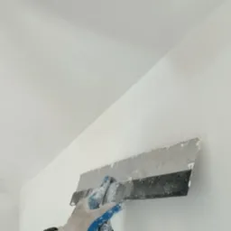 Nakładanie gładzi szpachlowej na ścianę przy suficie za pomocą szerokiej szpachelki przez osobę w rękawicy roboczej.