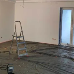 Pokój przed malowaniem: zabezpieczona folią podłoga, drabina malarska, ściany przygotowane do malowania, widoczne kable i puszki elektryczne.