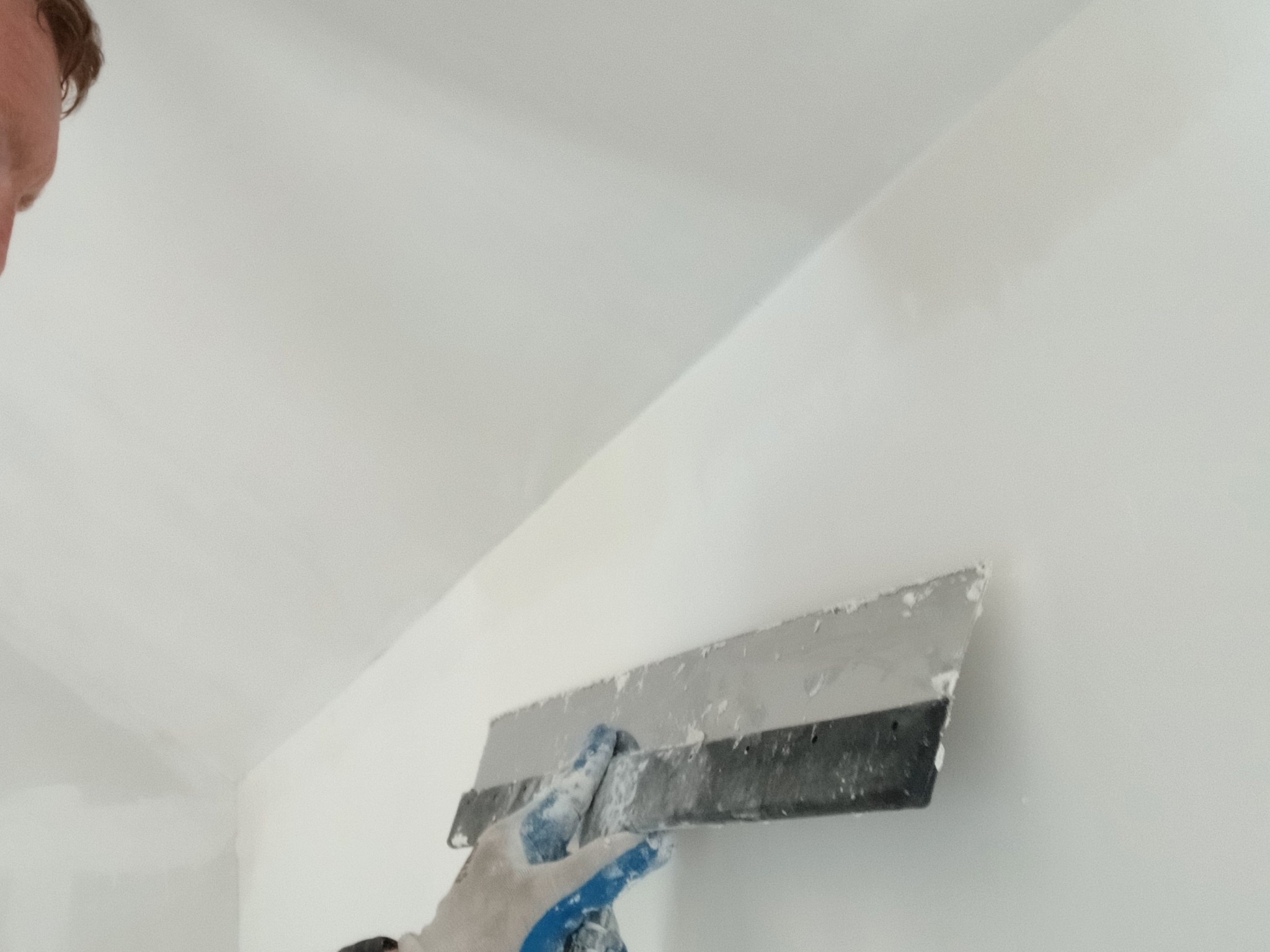 Nakładanie gładzi szpachlowej na ścianę przy suficie za pomocą szerokiej szpachelki przez osobę w rękawicy roboczej.
