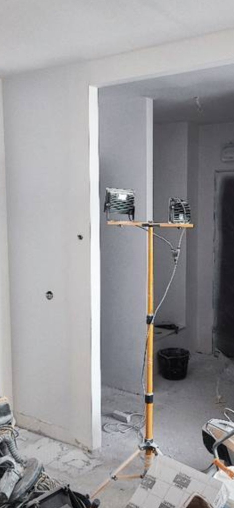 Wnętrze w trakcie remontu: surowe ściany, widoczne ślady po instalacjach elektrycznych, żółty statyw z dwoma reflektorami halogenowymi, wiadro i narzędzia budowlane na podłodze.