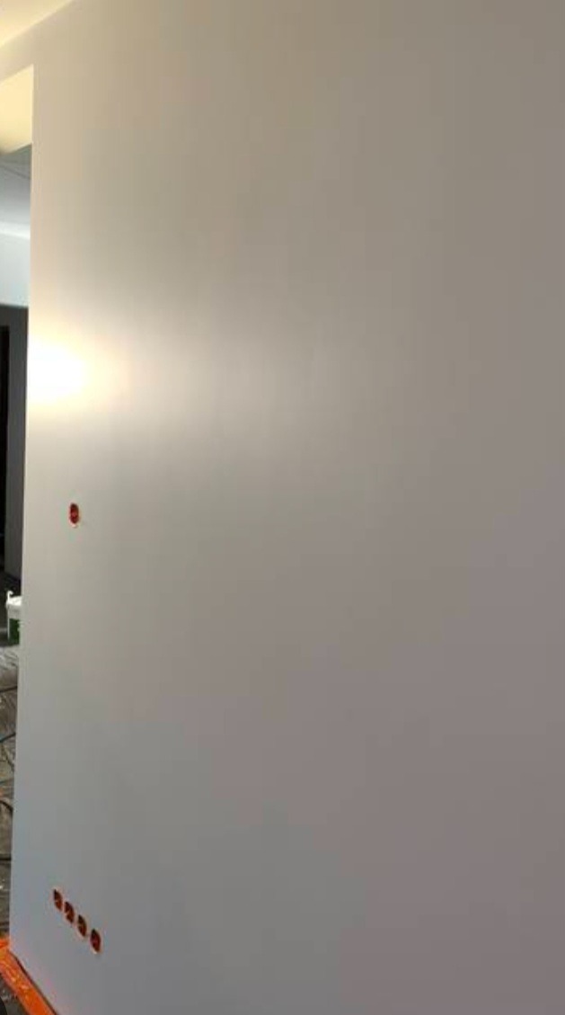 Świeżo pomalowana, gładka, biała ściana z widocznymi otworami po gniazdkach elektrycznych, zabezpieczonymi pomarańczową taśmą, w tle widoczny fragment wnętrza.