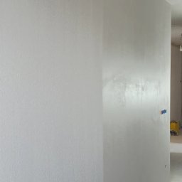 Mar-Gips - Wykończona, jasnoszara ściana z widoczną fakturą gładzi, z zamontowanymi, niepodłączonymi jeszcze, niebiesko-czerwonymi zaworami instalacji wodnej i śladami po szpachlowaniu.