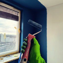 Mar-Gips - Malowanie ściany na granatowo wałkiem Anza trzymanym w zielonej rękawicy ochronnej; widoczne okno z zabezpieczeniami i drabina.