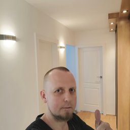 Mar-Gips - Mężczyzna z krótką brodą i włosami, ubrany w czarną koszulkę z plamami farby, pokazuje kciuk w górę na tle świeżo pomalowanego korytarza z dwoma białymi drzwiami, nowoczesnymi lampami ściennymi...