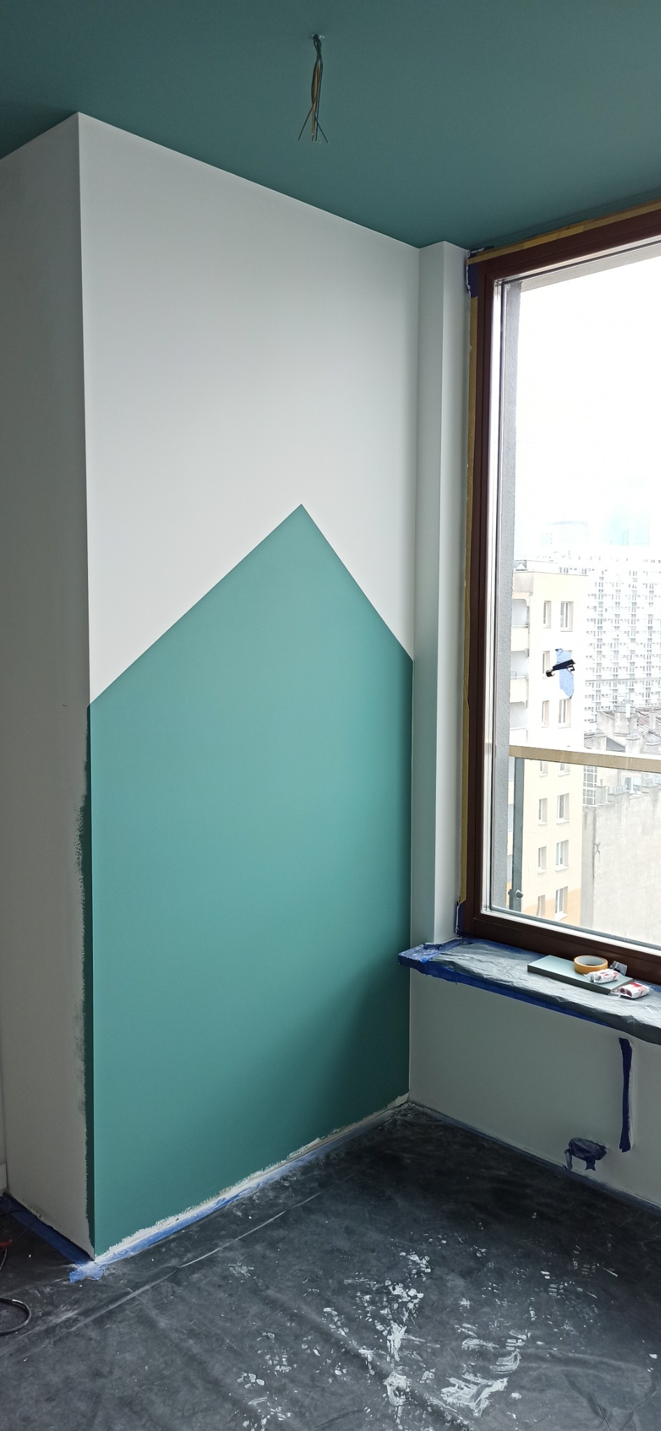 Pokój w trakcie malowania: ściana z geometrycznym podziałem kolorów - biały u góry, turkusowy trójkąt u dołu, okno z widokiem na miasto, podłoga zabezpieczona folią malarską, taśma malarska przy...