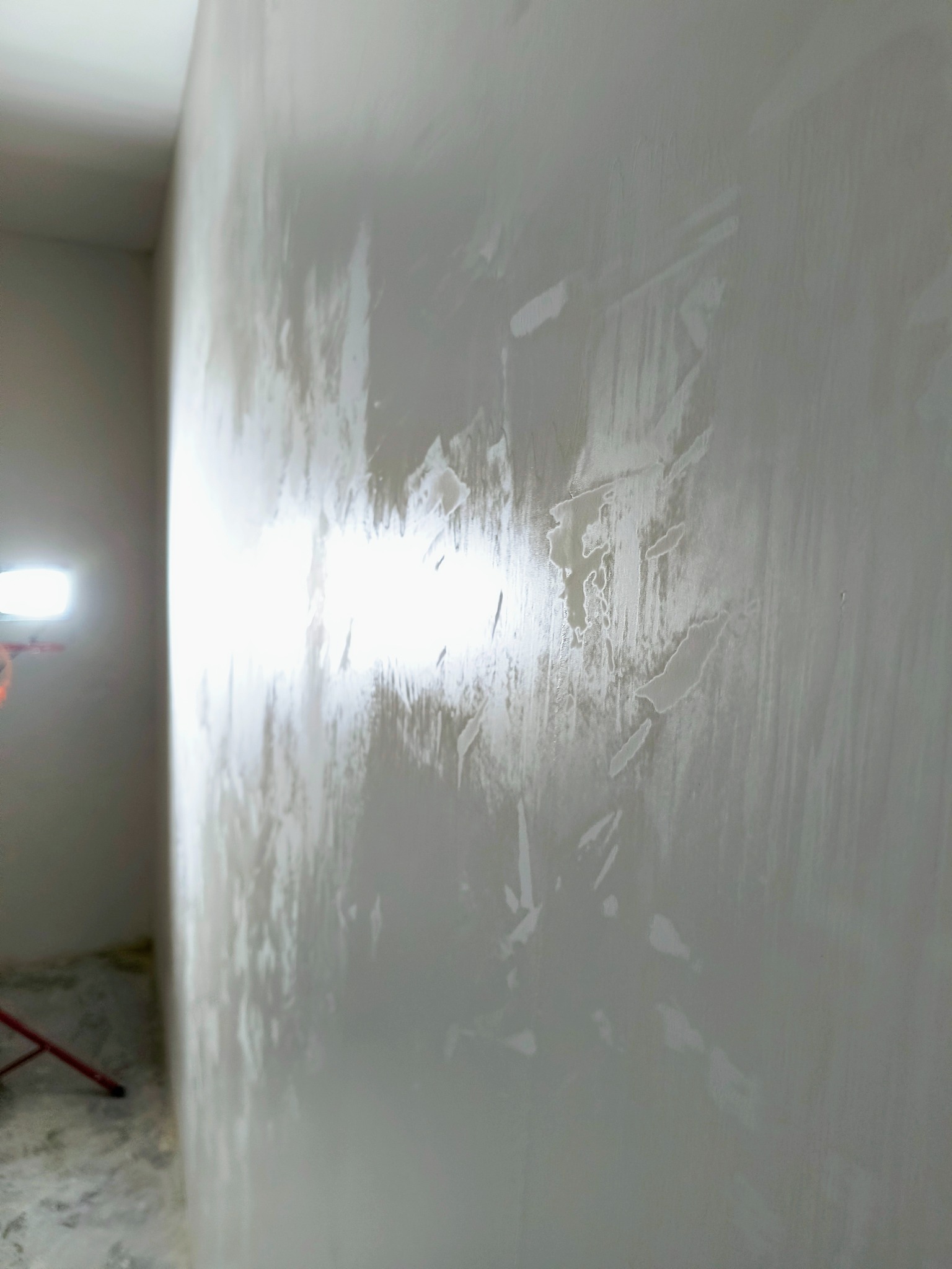 Ściana w trakcie szpachlowania, widoczne niedokładności i nierówności, oświetlona lampą roboczą.