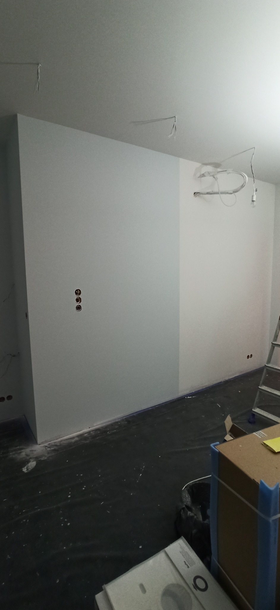 Świeżo pomalowane, jasnoszare ściany w trakcie remontu, z widocznymi przewodami elektrycznymi wystającymi z sufitu, zabezpieczoną folią podłogą i kartonami w rogu pomieszczenia.