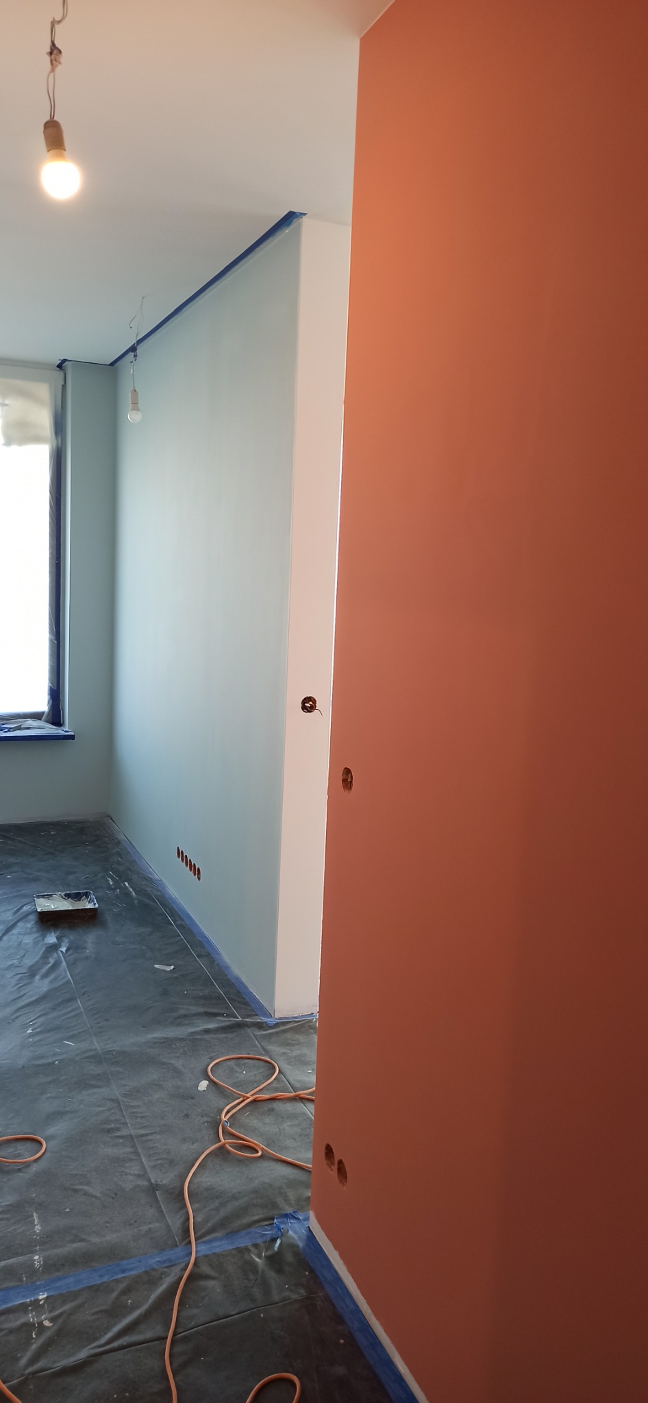 Pokój po świeżym pomalowaniu ścian na dwa kolory: łososiowy i jasnoniebieski, zabezpieczony folią malarską na podłodze i taśmą malarską przy suficie, widoczne puszki elektryczne bez osprzętu.