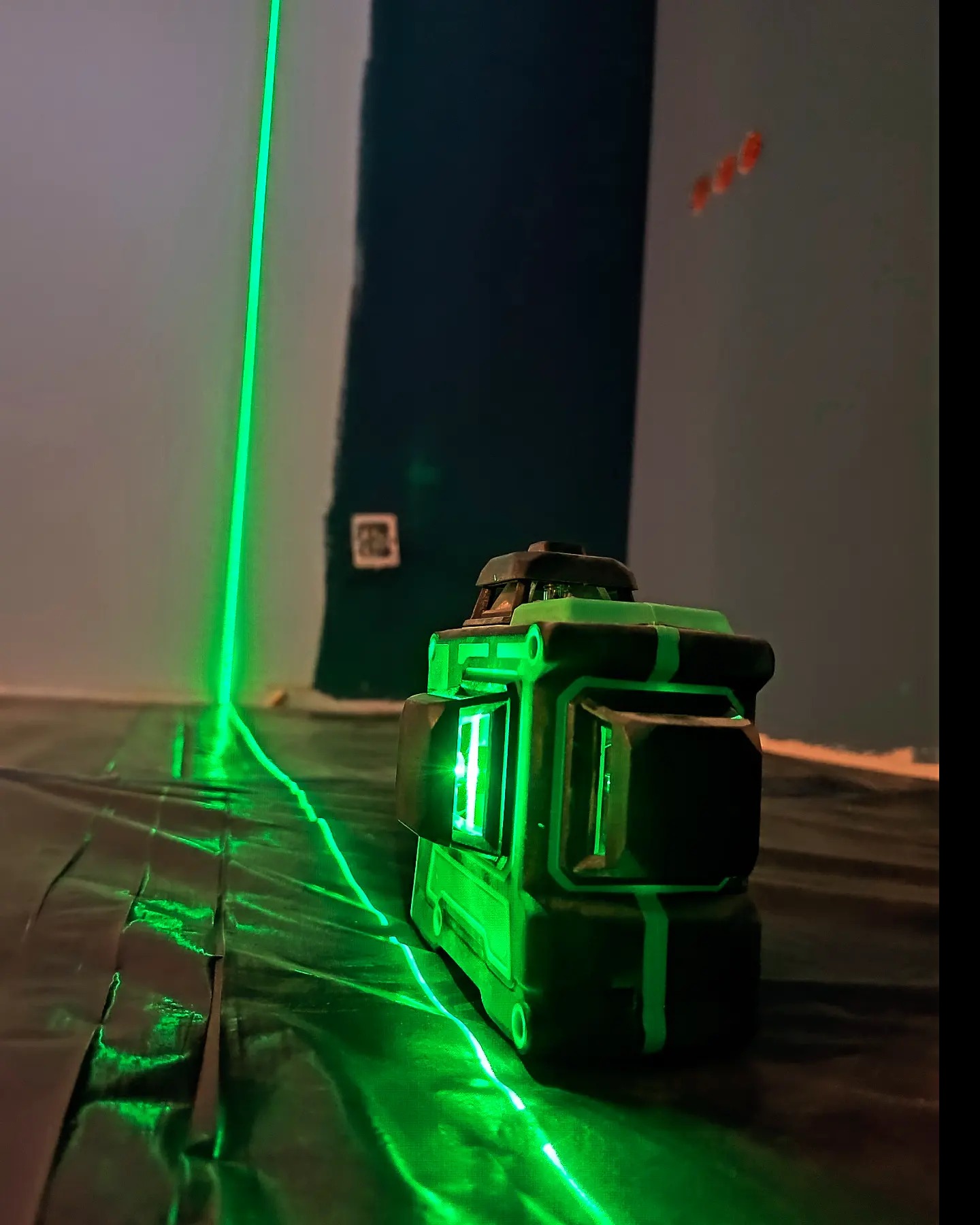 Zielony laser krzyżowy wyświetlający linie na ścianie i podłodze zabezpieczonej folią malarską, widoczne ślady po szpachlowaniu na ścianie.