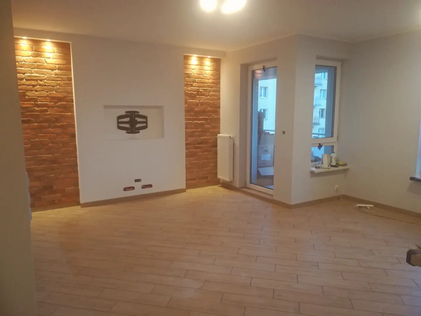 Salon po remoncie z ceglaną ścianą oświetloną od góry, drewnopodobną podłogą i przygotowanym miejscem na telewizor.