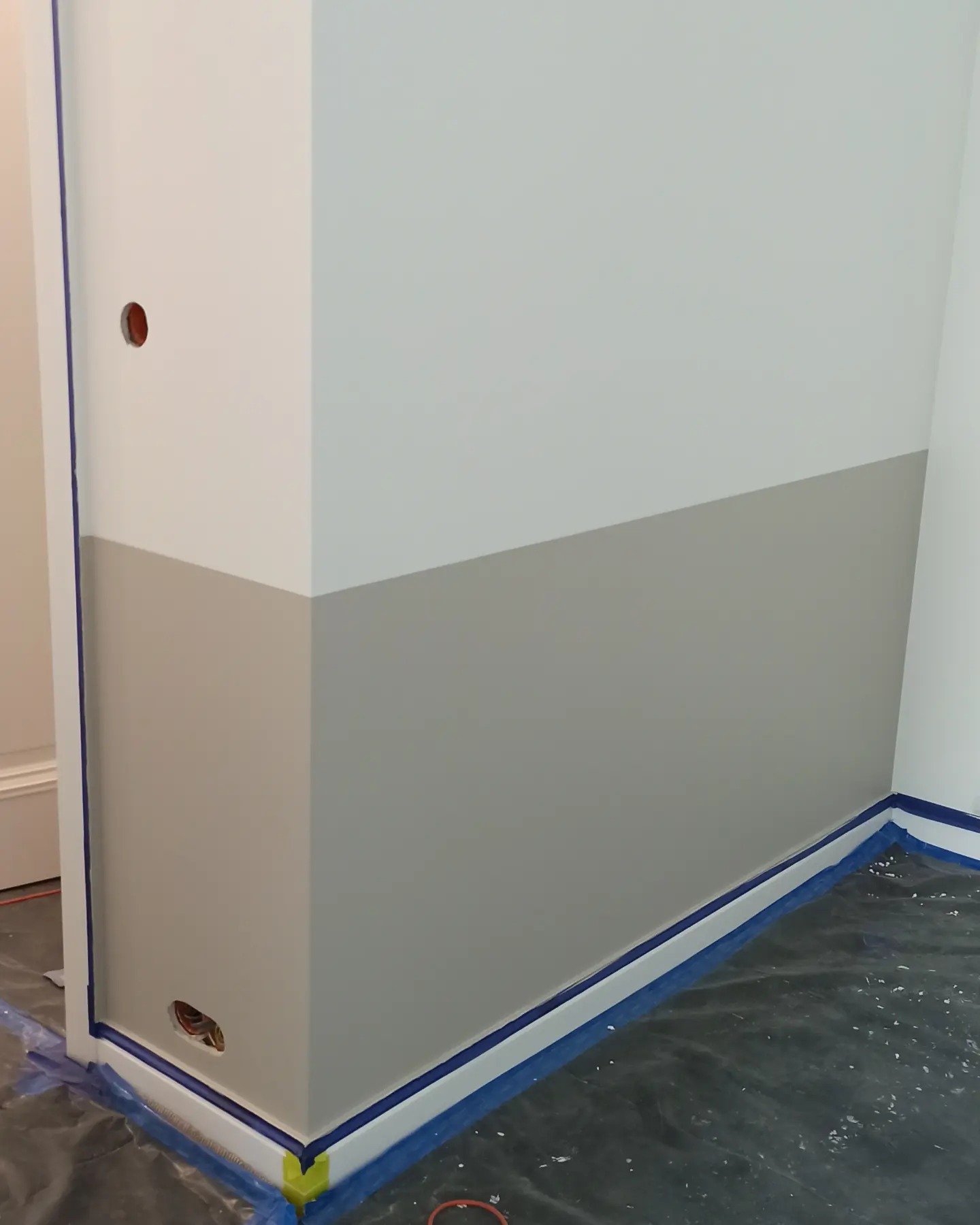 Świeżo pomalowany fragment ściany na dwa kolory, z zabezpieczonymi listwami przypodłogowymi niebieską taśmą malarską i folią ochronną na podłodze.