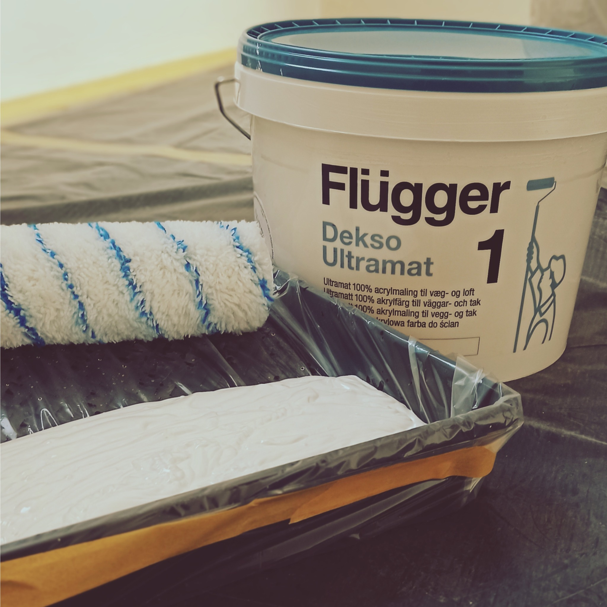 Pędzel wałkowy z niebieskimi paskami leżący obok kuwety z białą farbą i wiadra z farbą Flugger Dekso Ultramat 1 na folii ochronnej.