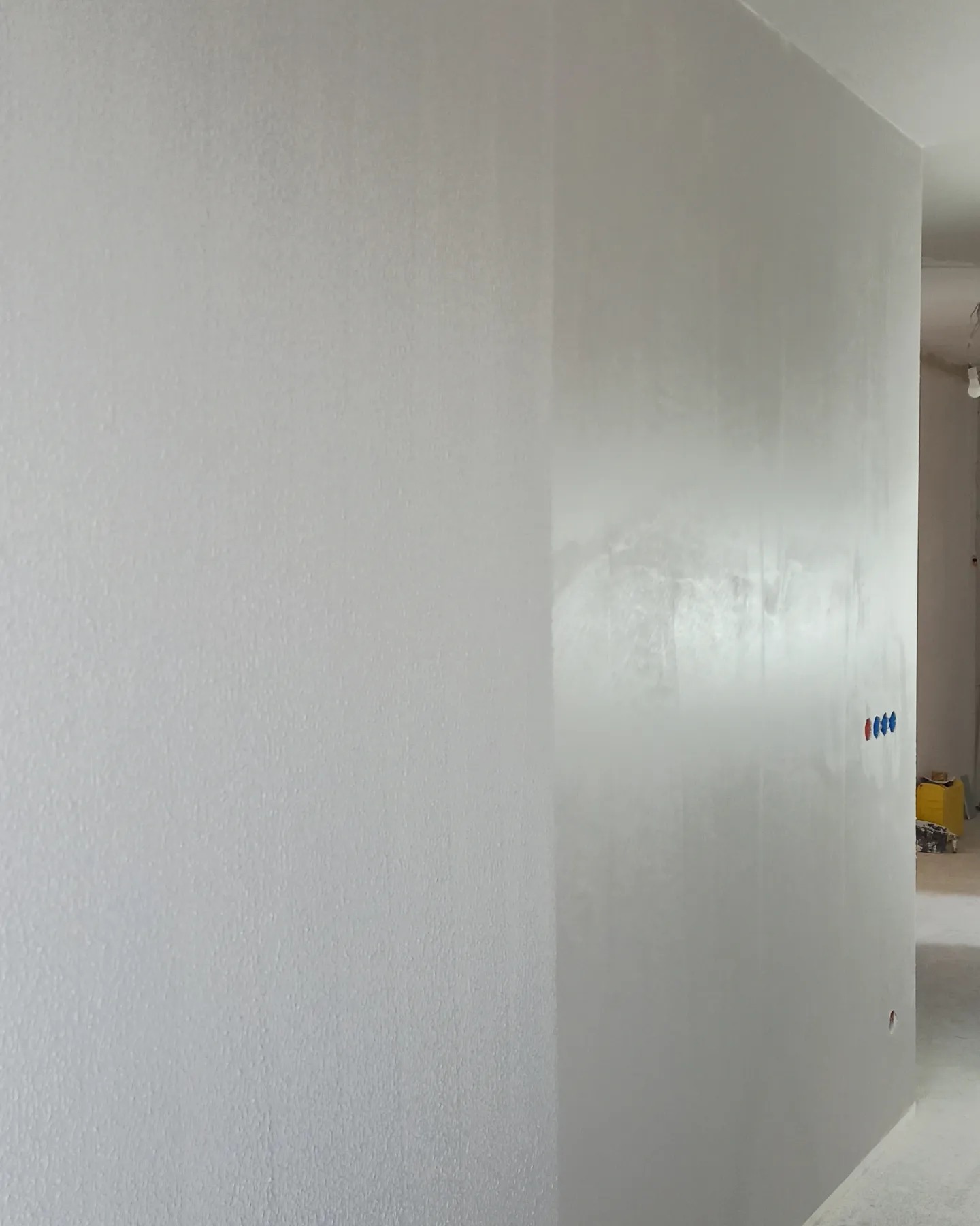 Wykończona, jasnoszara ściana z widoczną fakturą gładzi, z zamontowanymi, niepodłączonymi jeszcze, niebiesko-czerwonymi zaworami instalacji wodnej i śladami po szpachlowaniu.