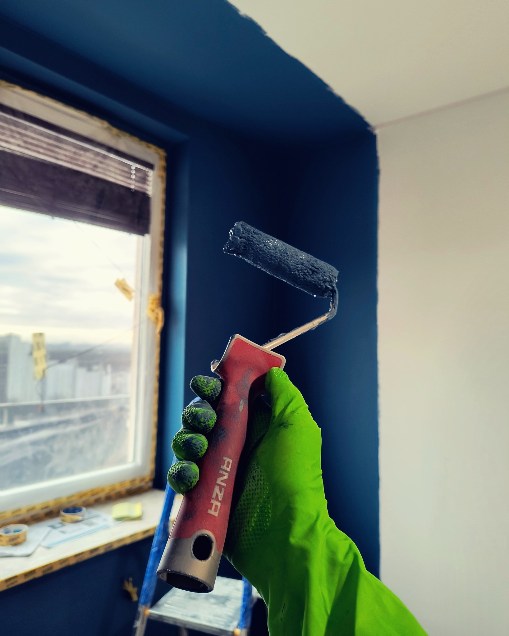 Malowanie ściany na granatowo wałkiem Anza trzymanym w zielonej rękawicy ochronnej; widoczne okno z zabezpieczeniami i drabina.