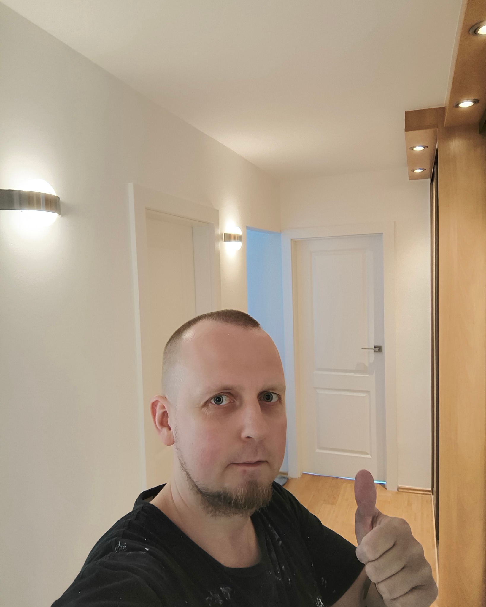 Mężczyzna z krótką brodą i włosami, ubrany w czarną koszulkę z plamami farby, pokazuje kciuk w górę na tle świeżo pomalowanego korytarza z dwoma białymi drzwiami, nowoczesnymi lampami ściennymi...