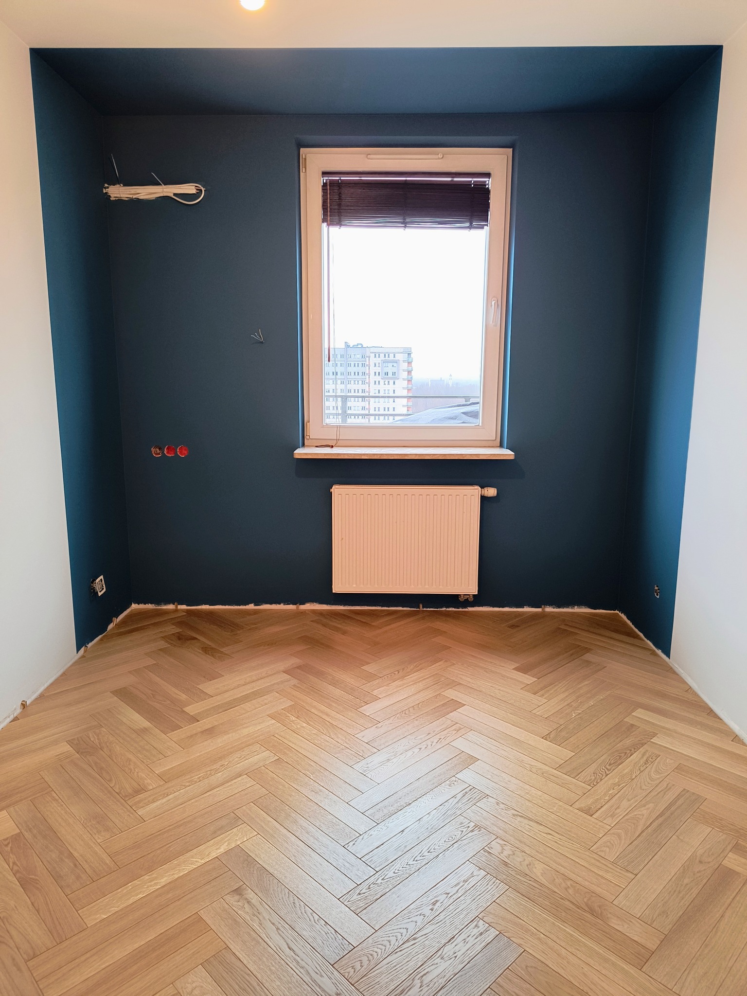 Pokój po remoncie z ciemnoniebieskimi ścianami, jasnym parkietem ułożonym w jodełkę, białymi narożnikami i grzejnikiem pod oknem. Widoczne niedokończone instalacje elektryczne.