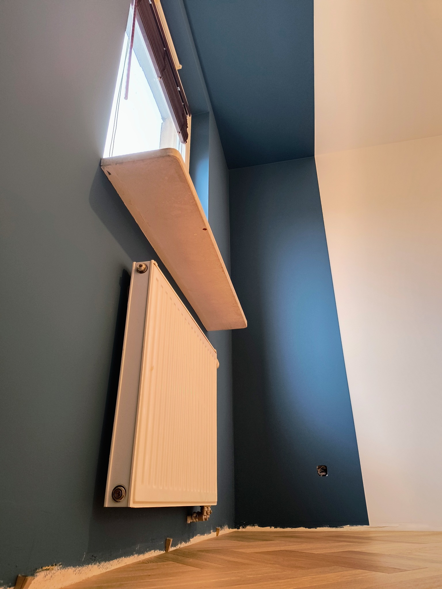 Pomieszczenie po malowaniu ścian na ciemny niebieski kolor, widoczny grzejnik, parapet i fragment podłogi z parkietem w jodełkę, listwy przypodłogowe jeszcze nie zamontowane.