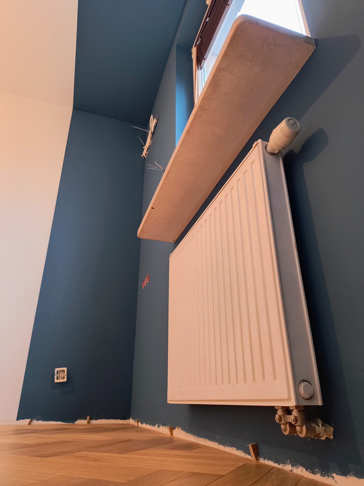 Ściana pomalowana na ciemny odcień niebieskiego, z widocznym grzejnikiem, parapetem z betonu architektonicznego i fragmentem parkietu ułożonego w jodełkę. Widoczne niedociągnięcia malarskie przy...