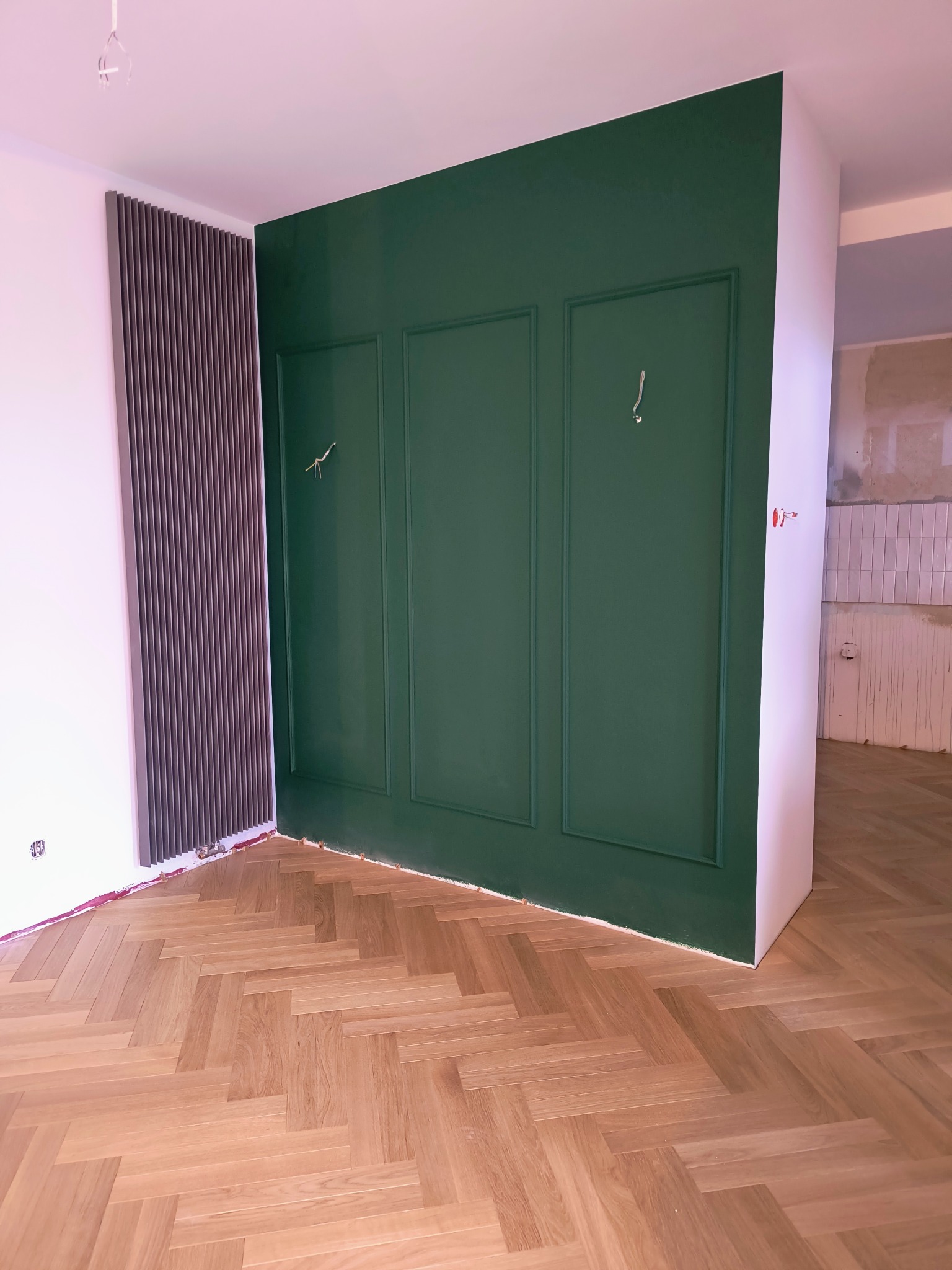 Świeżo pomalowana ściana w kolorze butelkowej zieleni z dekoracyjnymi listwami, widoczny parkiet w jodełkę i nowoczesny grzejnik panelowy, w tle fragment pomieszczenia w trakcie prac...