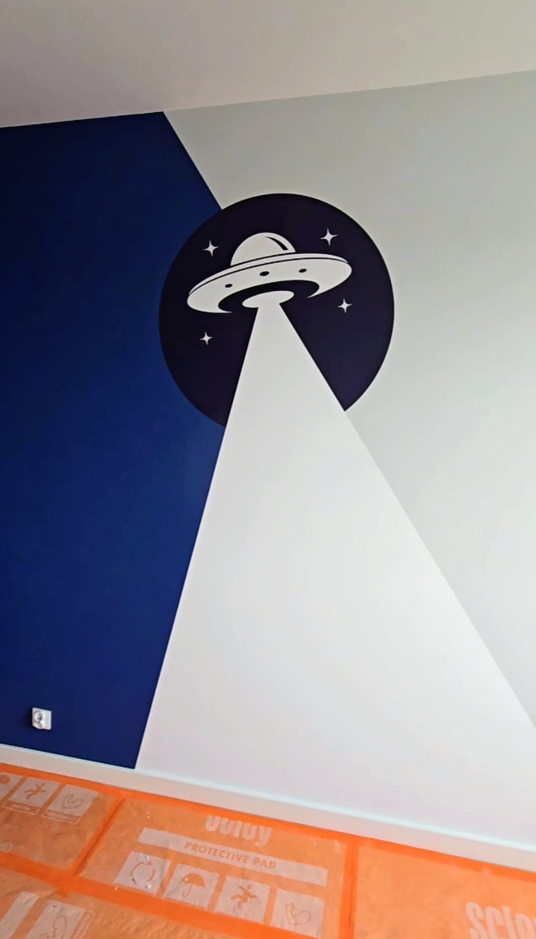 Ściana z motywem UFO w pokoju dziecięcym, pomalowana na biało i granatowo, z zabezpieczoną podłogą. Nietypowy wzór i kolorystyka.