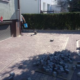 Sporco Poznań - Podjazd z częściowo ułożonej czerwono-brązowej kostki brukowej z jasnymi akcentami na łuku, obok stos szarych kostek i pracownik z niebieskim wiadrem.