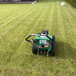 GRU GARDEN - Zielony aerator do trawy na trawniku, widoczne rdzenie wyciągnięte z gleby, w tle nowoczesny dom z tarasem i zadbany ogród.