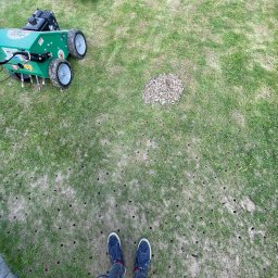 GRU GARDEN - Aeracja trawnika z widocznymi otworami i wyciągniętymi korkami ziemi, dwie kosiarki w tle, ujęcie z góry na stopy osoby wykonującej prace.