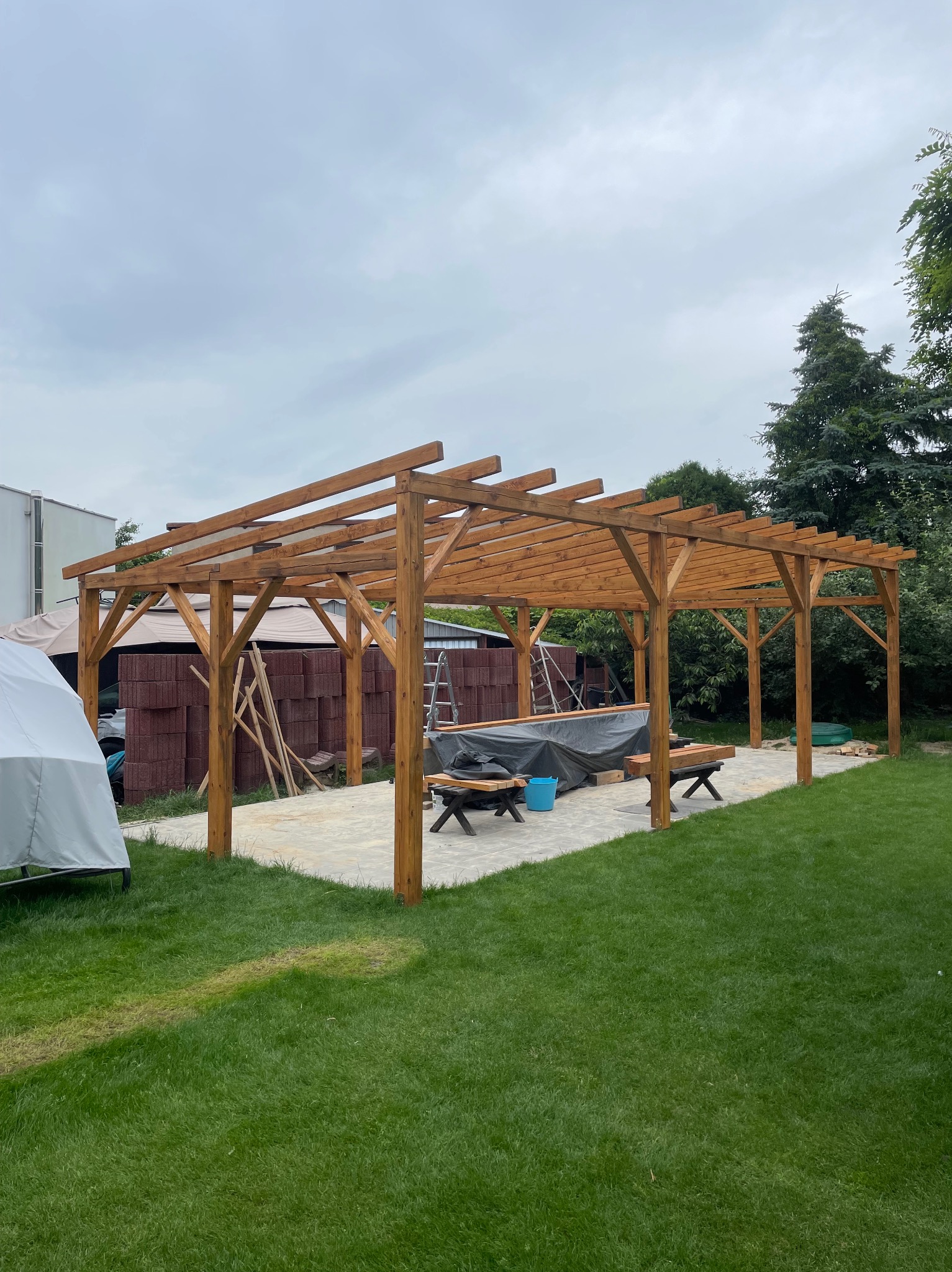 Drewniana pergola w trakcie budowy na trawniku, widoczne materiały budowlane i narzędzia, szara kostka brukowa, pochmurne niebo w tle.